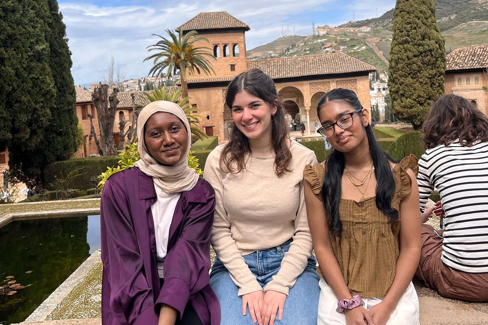 Three Jewish Studies students sit together on a low wall at the Alhambra in Granada, with the Partal palace and the Generalife gardens behind them.