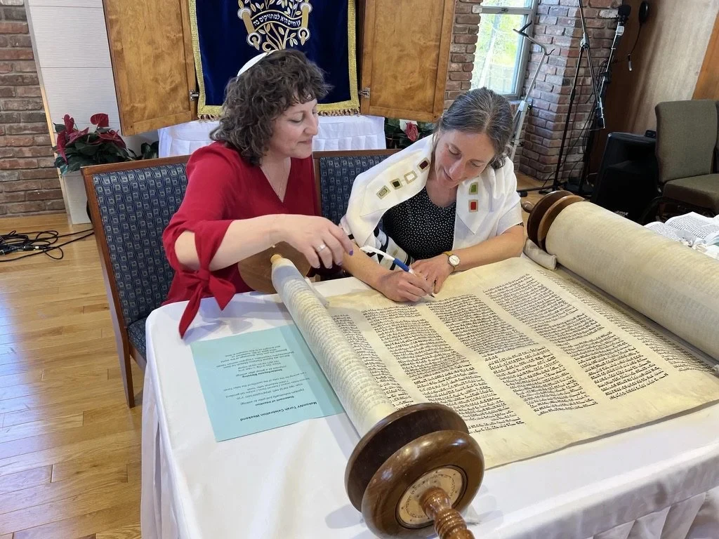 Julie Seltzer guides a congregant&rsquo;s hand on the quill at a public Torah-writing event; a Sefer Torah opened on a draped table in a synagogue sanctuary with an ark and embroidered parokhet visible behind them.