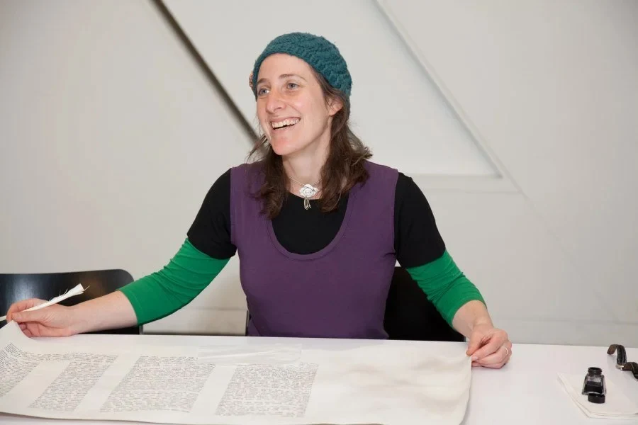 Julie Seltzer, soferet, seated at her writing table holding a feather quill, with a parchment scroll opened before her and an inkpot at her right hand.